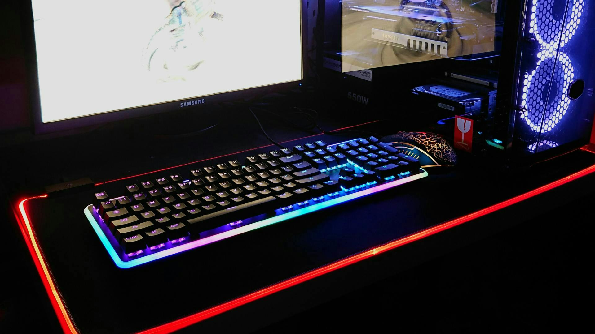 An LED illuminated gaming keyboard stands on a gaming table next to a gaming PC with LED components.