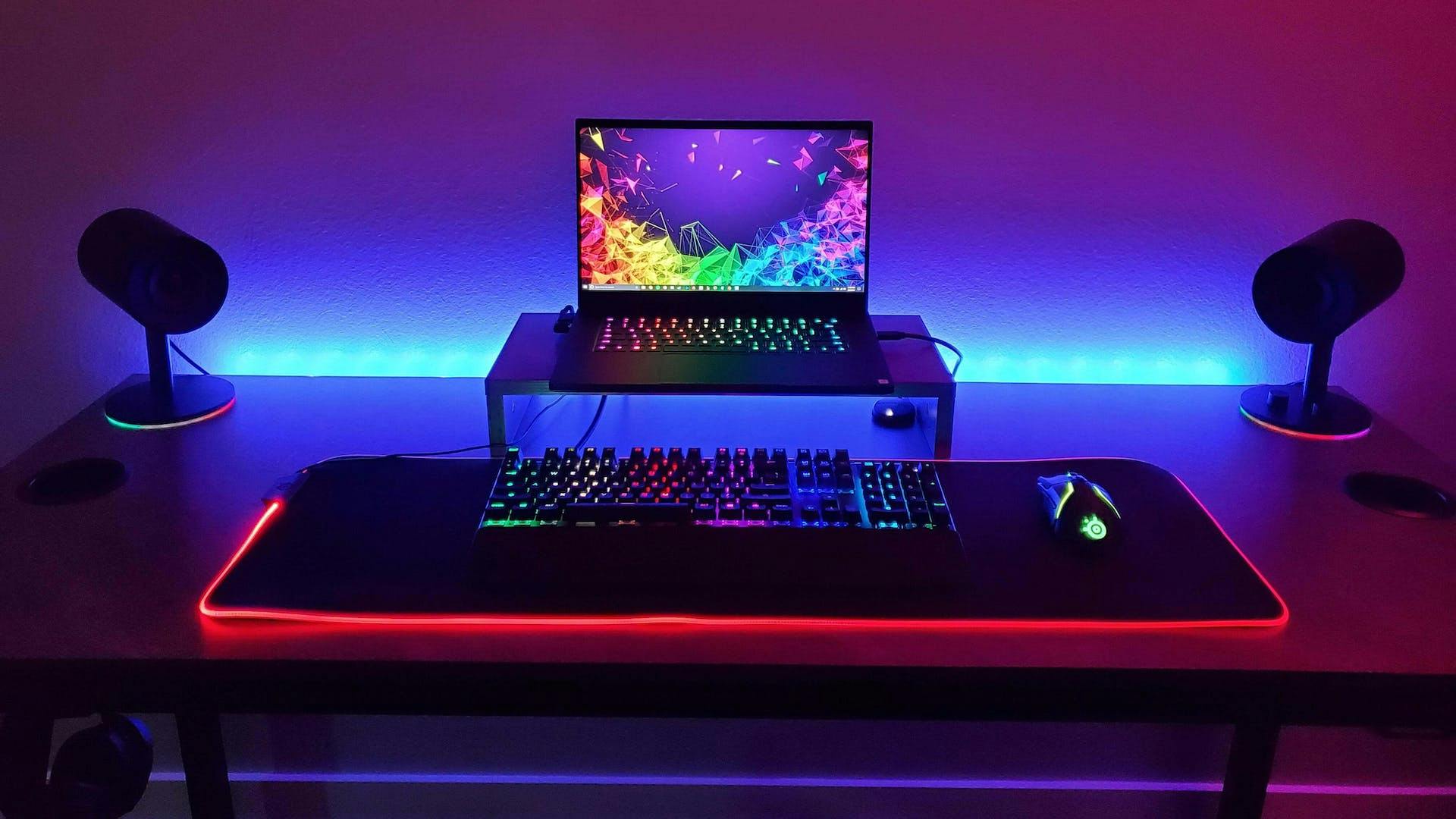 On a table without decoration but with many colorful gaming LED elements, a laptop stands frontally somewhat elevated behind a gaming keyboard.