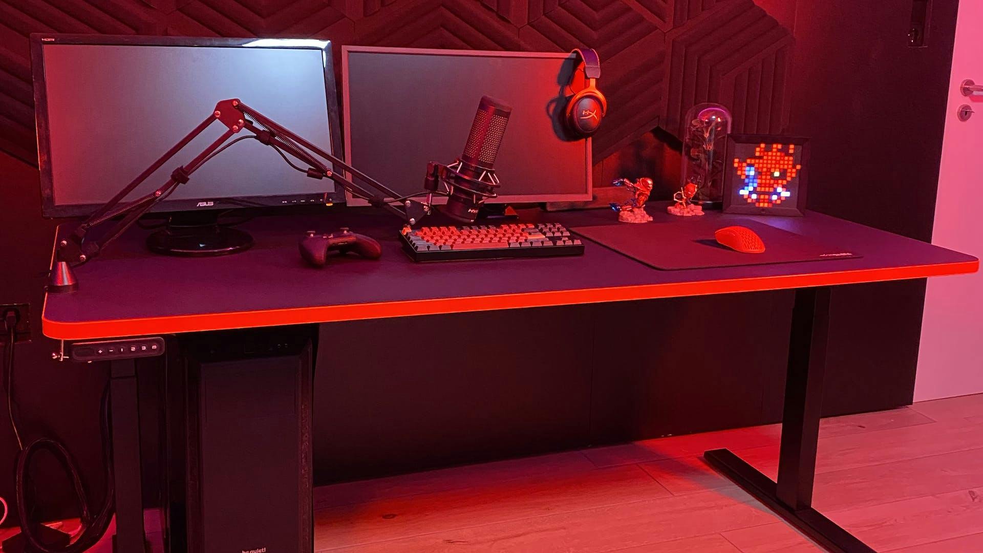 In einem rot leuchtenden Raum steht relevantes Gaming Setup Zubehör inklusive Mikrofon-Arm und Gaming Dekoration auf einem LeetDesk Gaming Tisch
