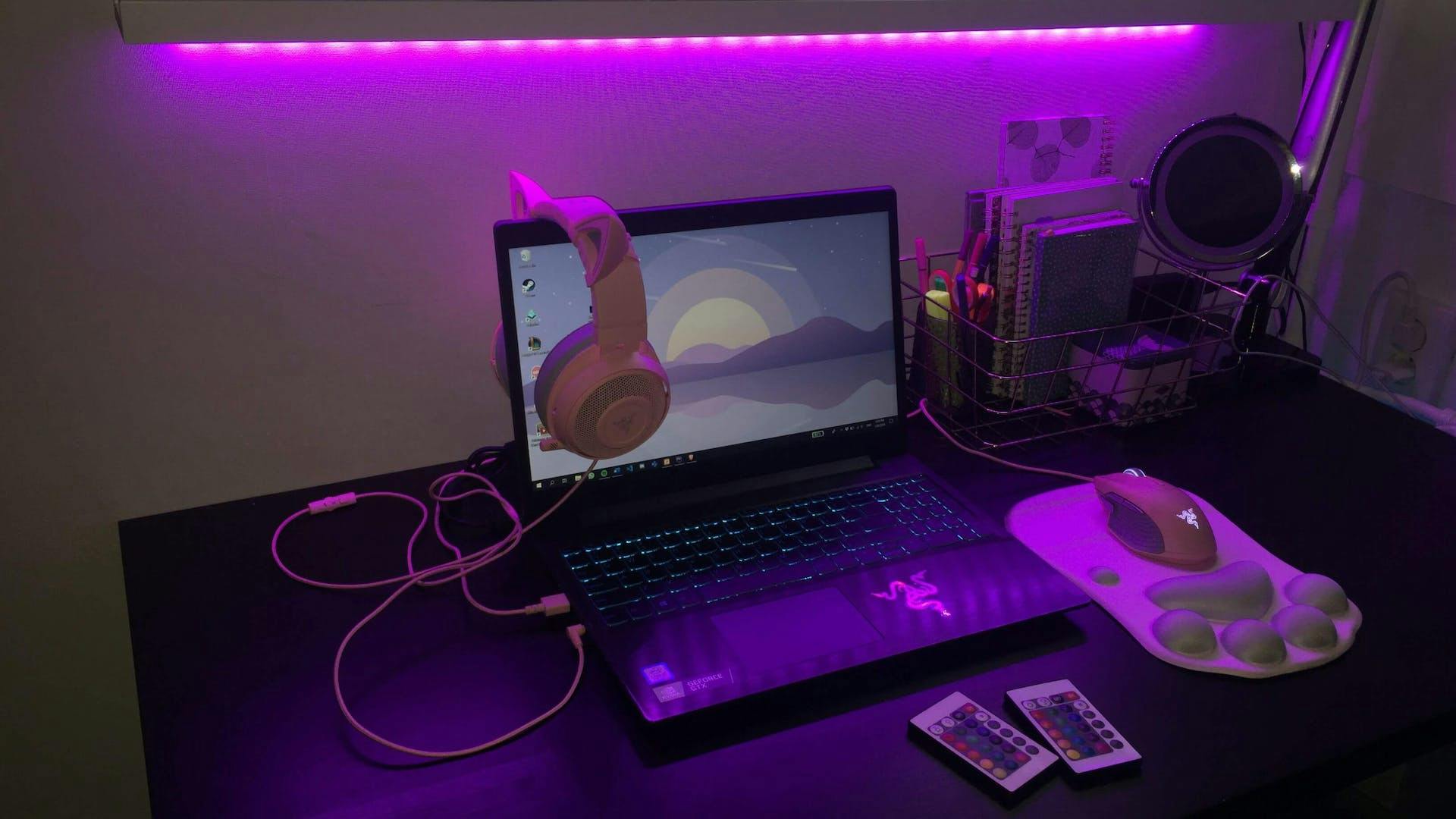 On a desk in a purple LED lit gaming room is a gaming laptop, gaming mouse and a matching gaming headset over the screen