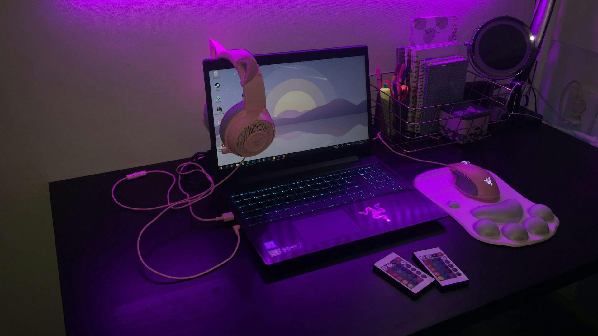 Auf einem Schreibtisch in einem lila LED beleuchteten Gaming Zimmer steht ein Gaming Laptop, Gaming Maus und einem farblich passenden Gaming Headset über dem Bildschirm