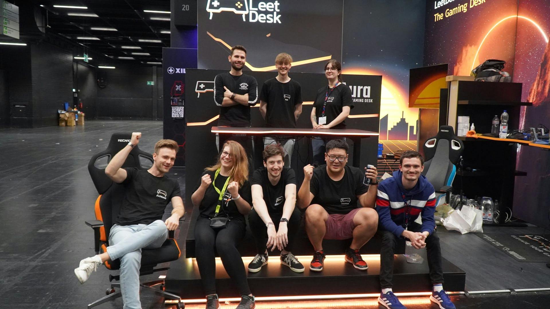 The LeetDesk Team is sitting on their Gamescom stange in front of and behind the AURA LED gaming desk, everyone is smiling.
