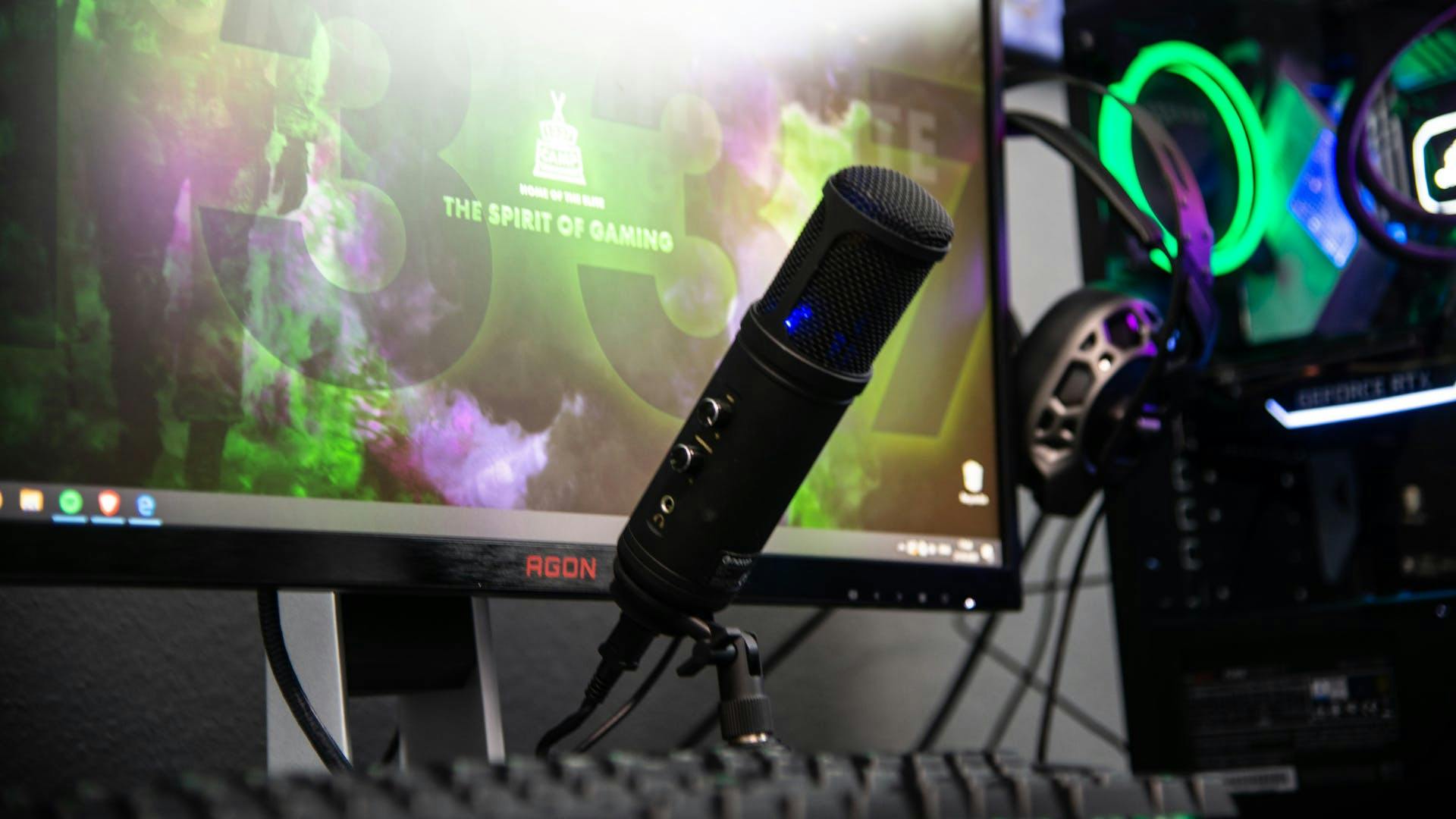 A professional microphone on a boom arm in front of a gaming monitor and an LED gaming PC