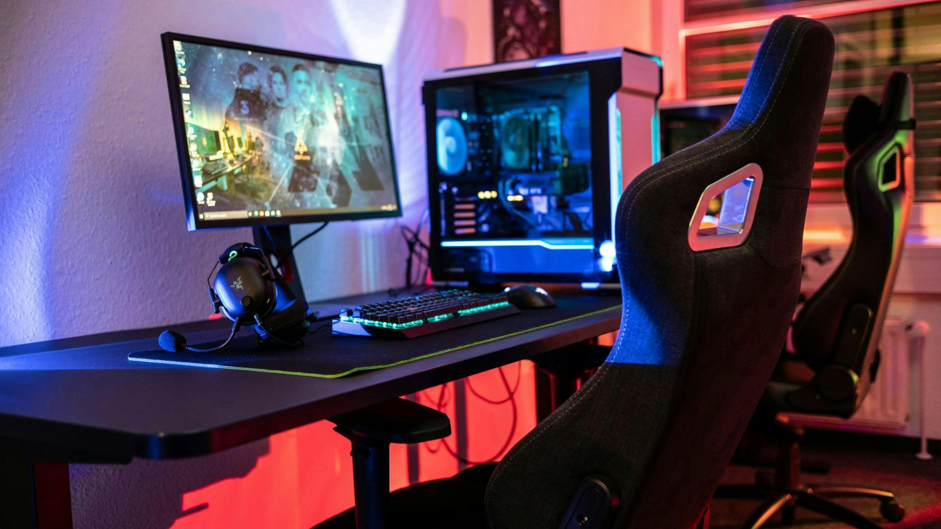 In einem LED beleuchteten Gaming Setup stehen zwei Gaming Stühle nebeneinander und blicken auf einen Gaming Monitor der zusammen mit einem Gaming PC und LED Tastatur auf einem Gaming Tisch steht