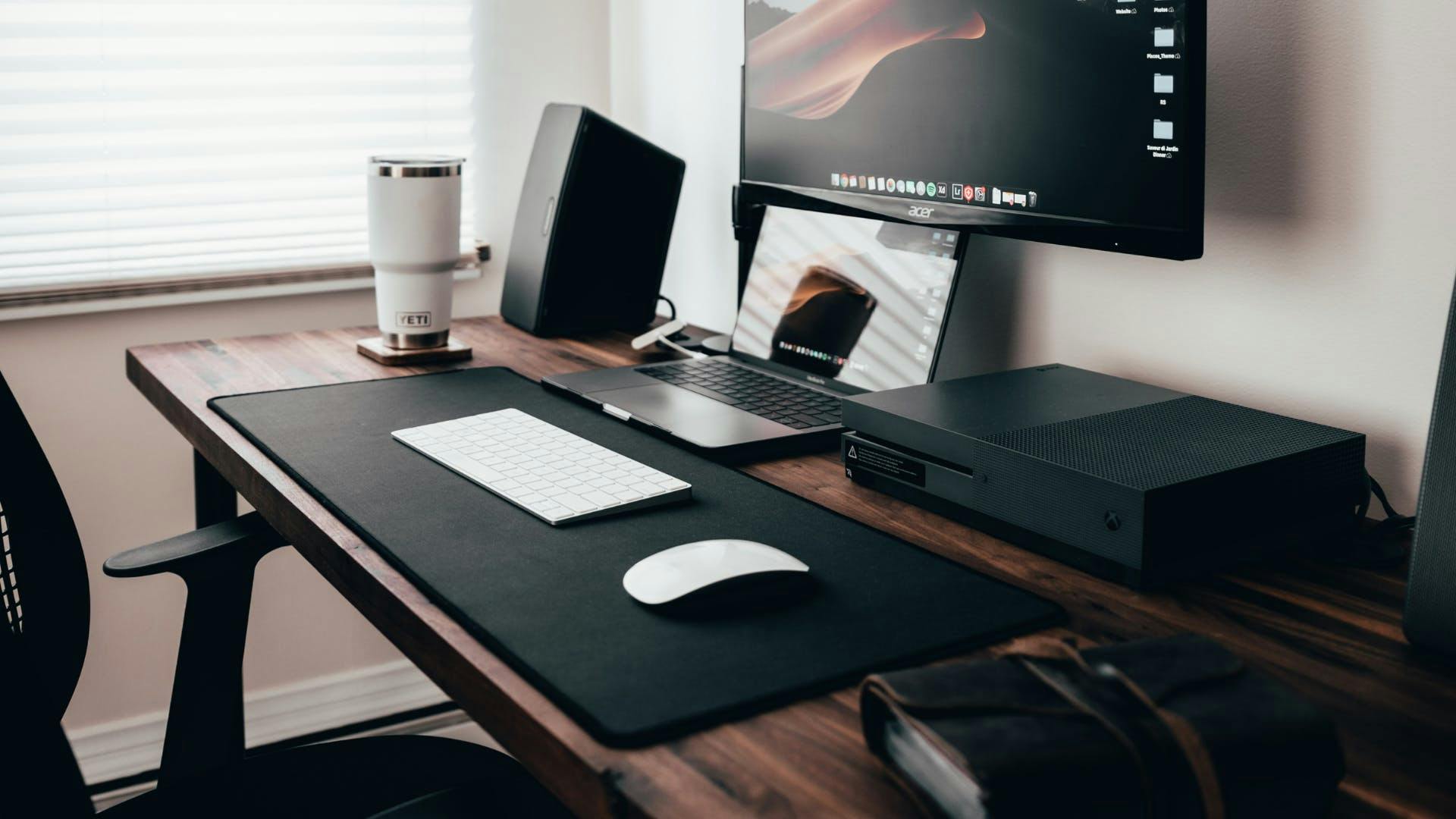Auf einem büro-ähnlichen, minimalistischen Schreibtisch in dunklen Holztönen steht ein Laptop und eine Tastatur vor einem Fernseh-Monitor und Lautsprechern