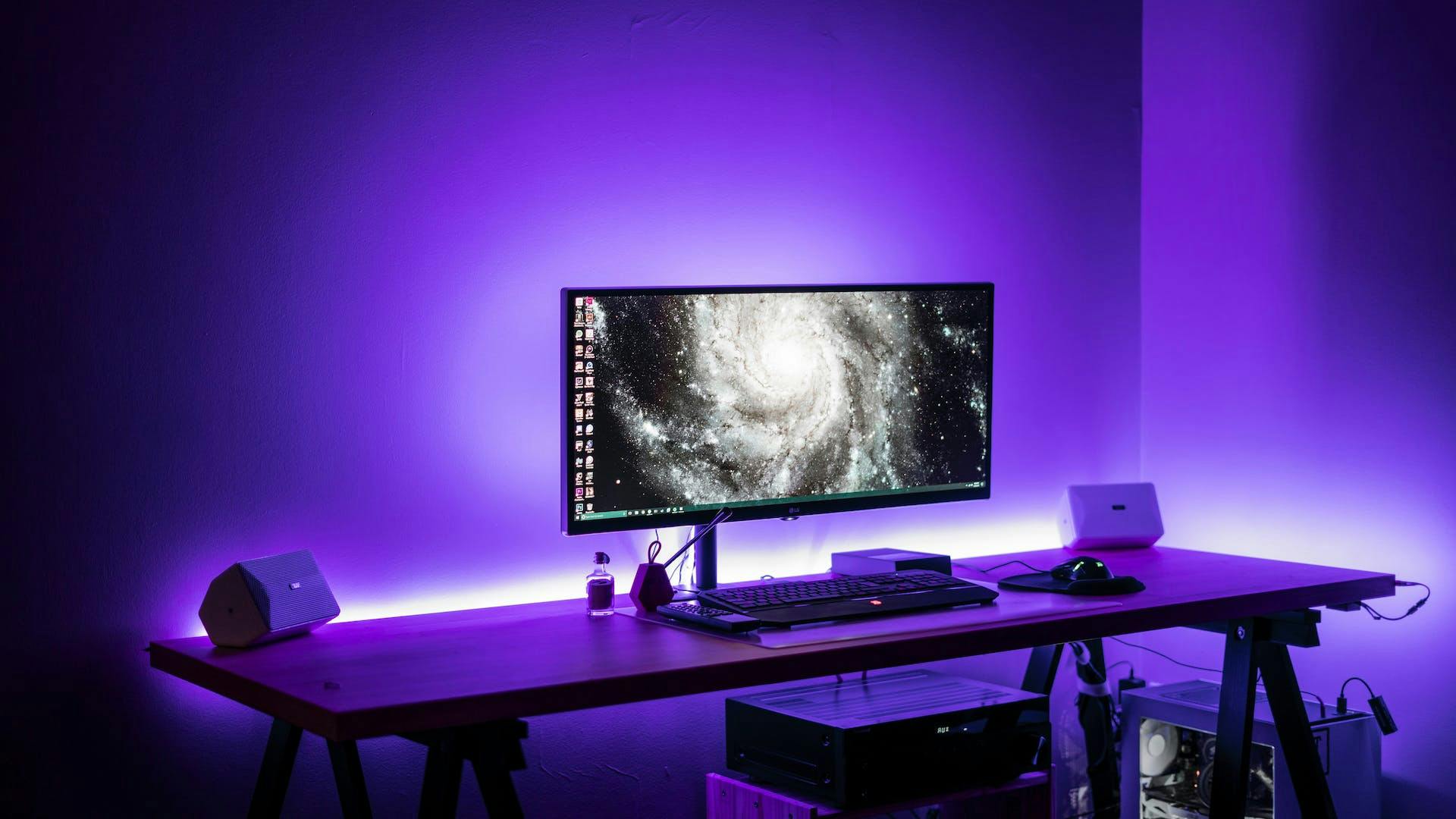 In einem clean gehaltenen Gaming Setup mit lila LED Elementen steht ein Gaming Monitor und eine Gaming Tastatur auf einem Gaming Tisch 
